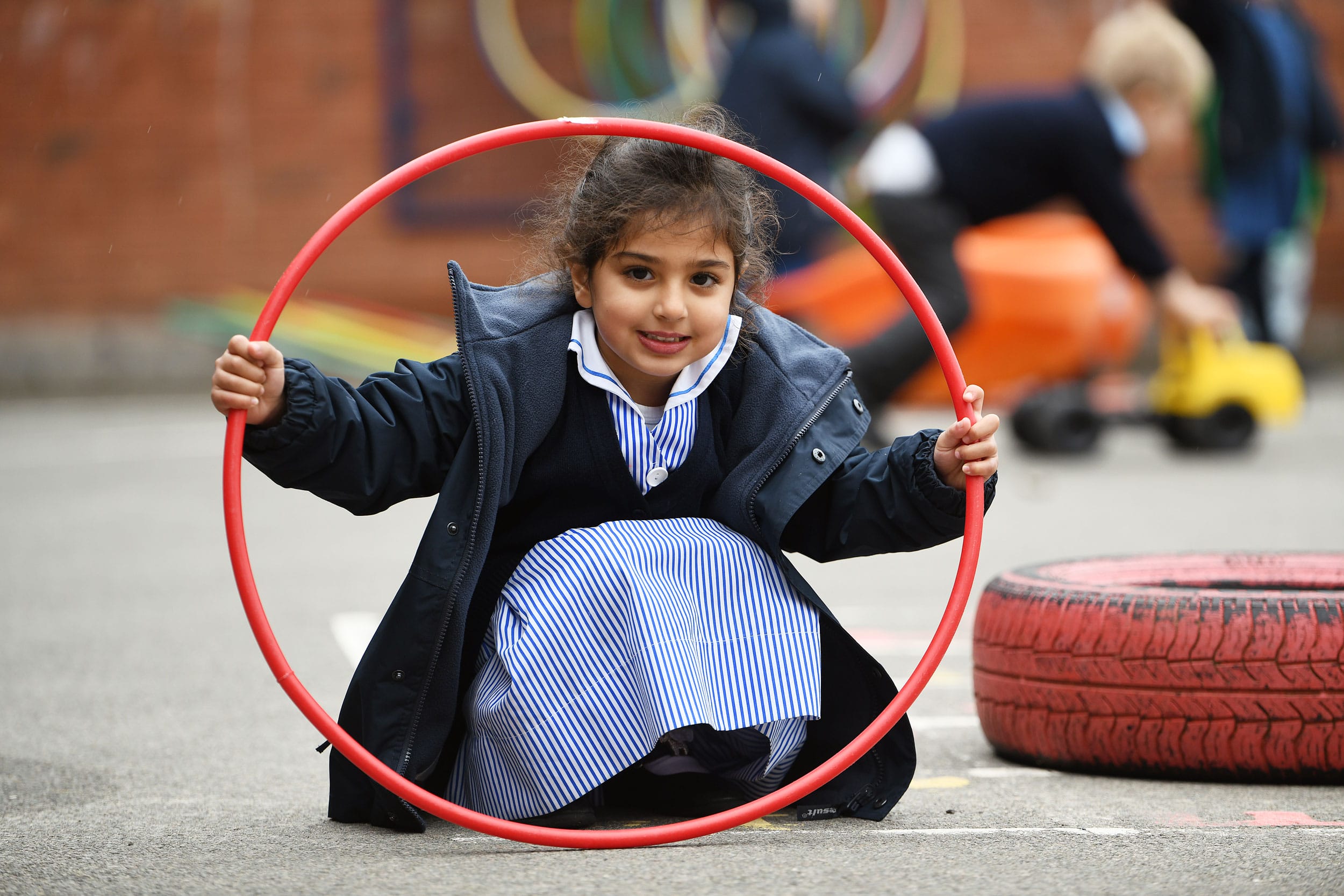 A Nursery girl crouched down and looking through a hula hoop on the playground.
