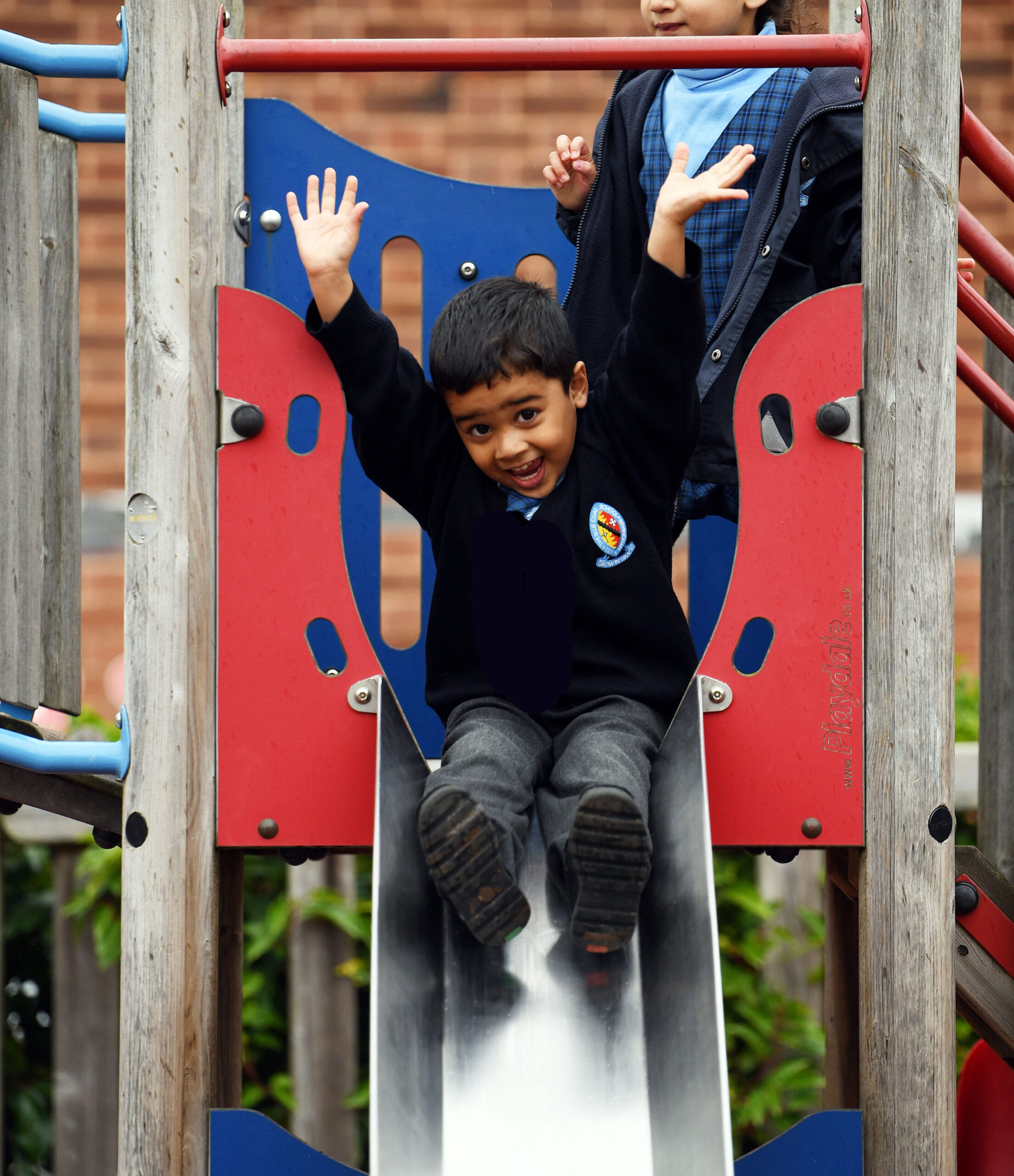 A boy slides down the slide in the Nursery playground with his arms in the air.