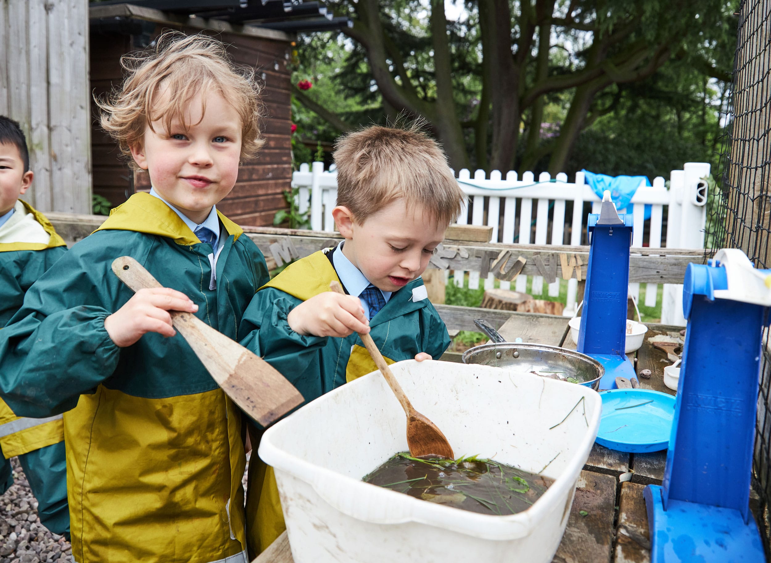 Two boys playing with a bowl of muddy water and wooden spoons in the Mud Kitchen in Forest School.