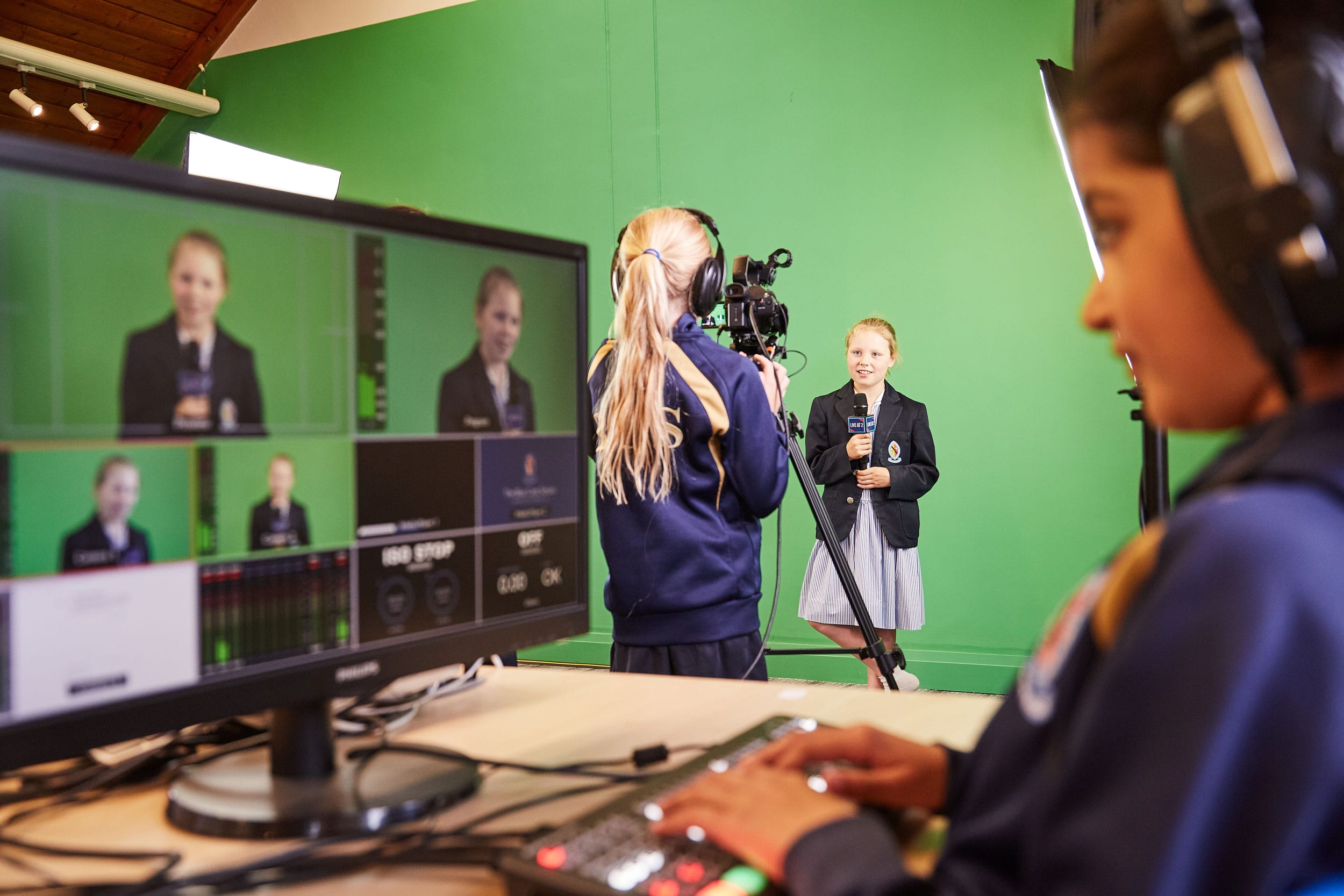 A pupil is being interviewed by another pupil behind a camera on a green screen and another pupil is looking at the editing screen.