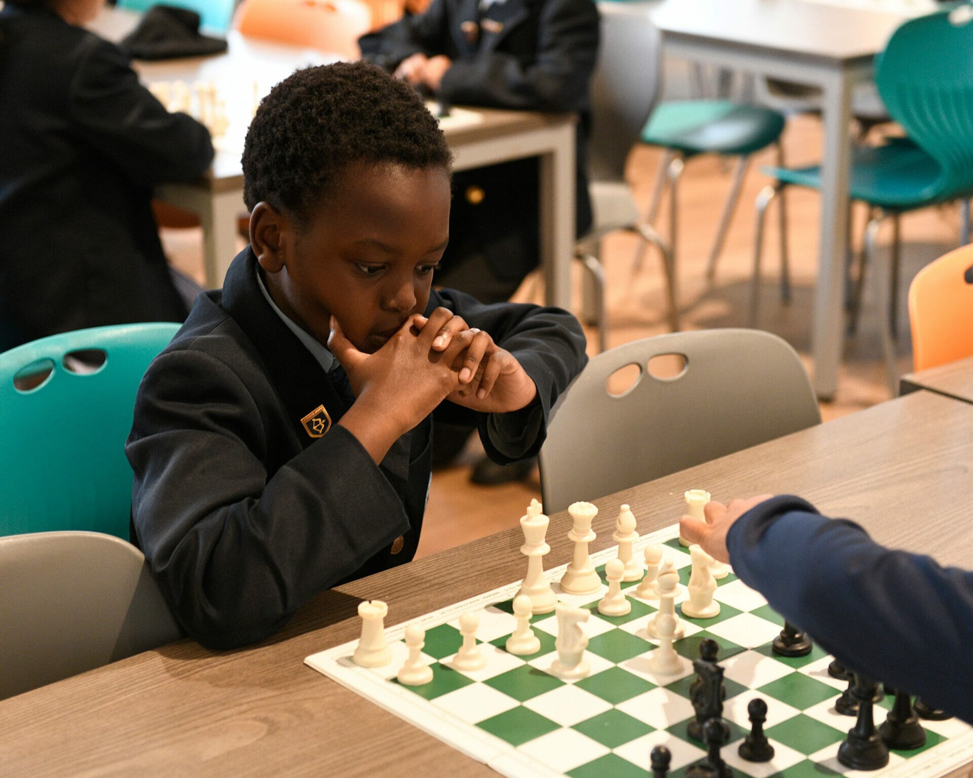 A boy is sat at a table playing chess. His opponent is making a move and the player in the photo is thinking.