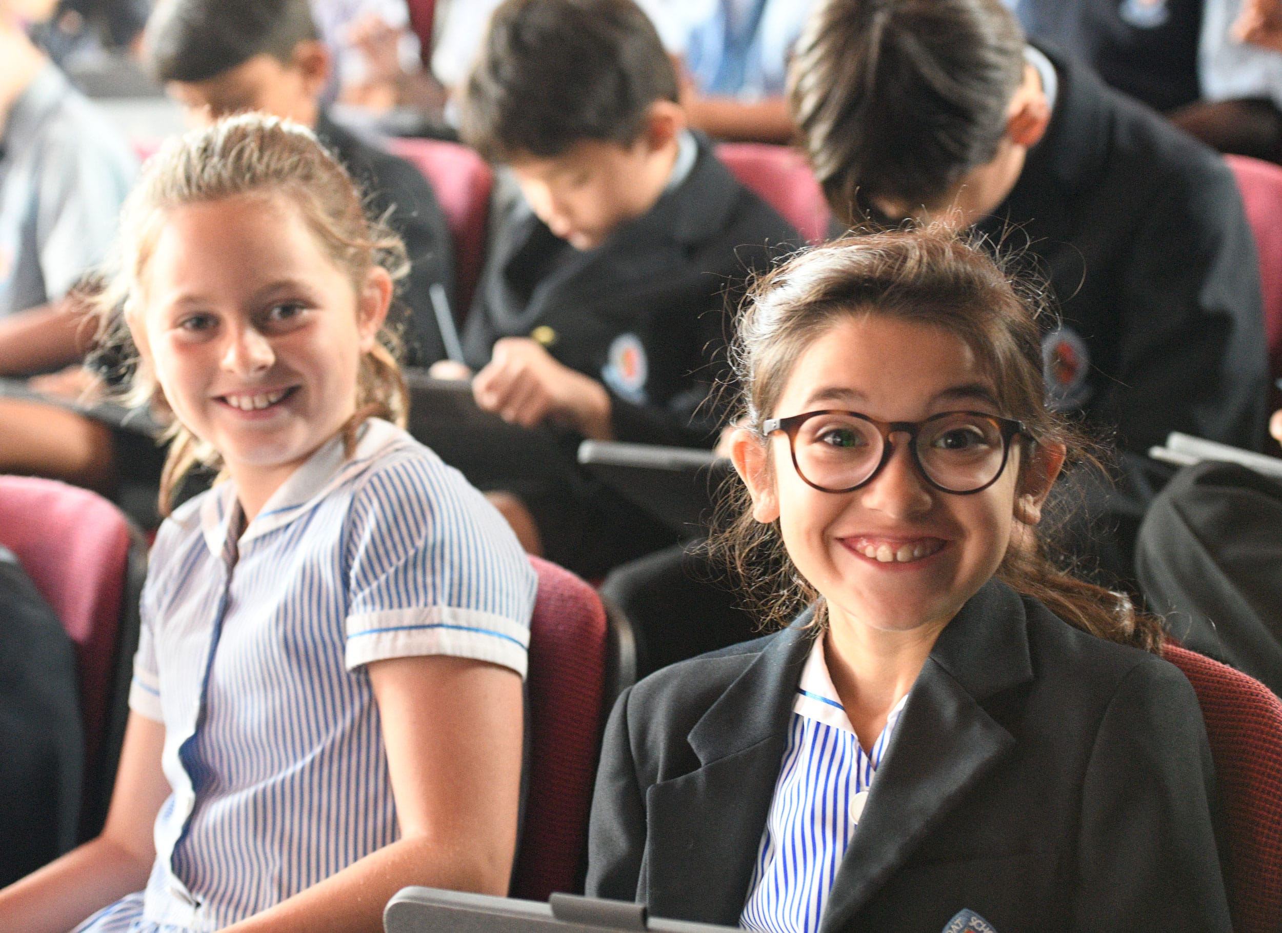 Two girls sat in the School Hall smile at the camera.