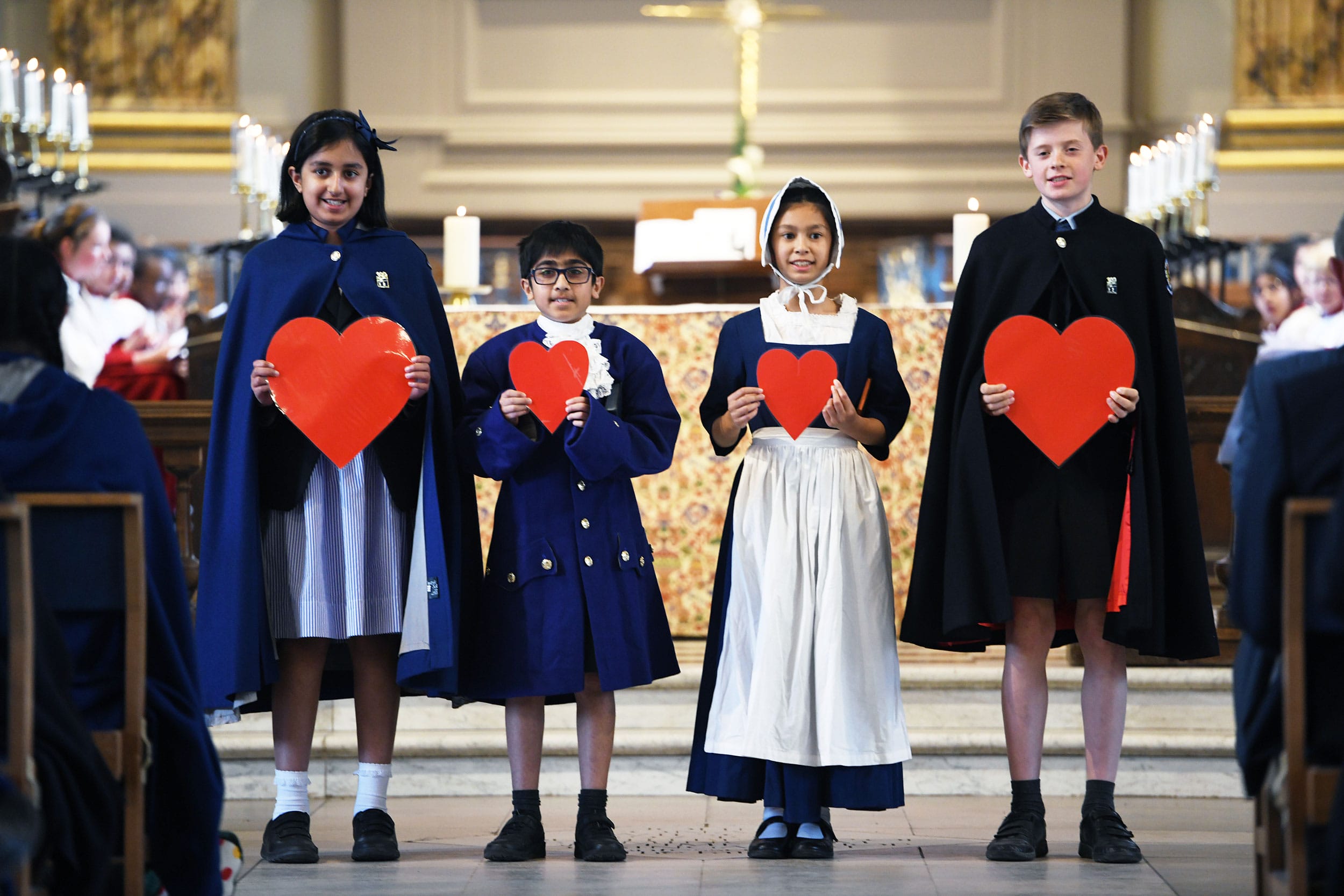 Four pupils stand in St Philips' Cathedral dressed in different outfits representing the School's history, holding paper hearts to symbolise the Blue Coat Values.