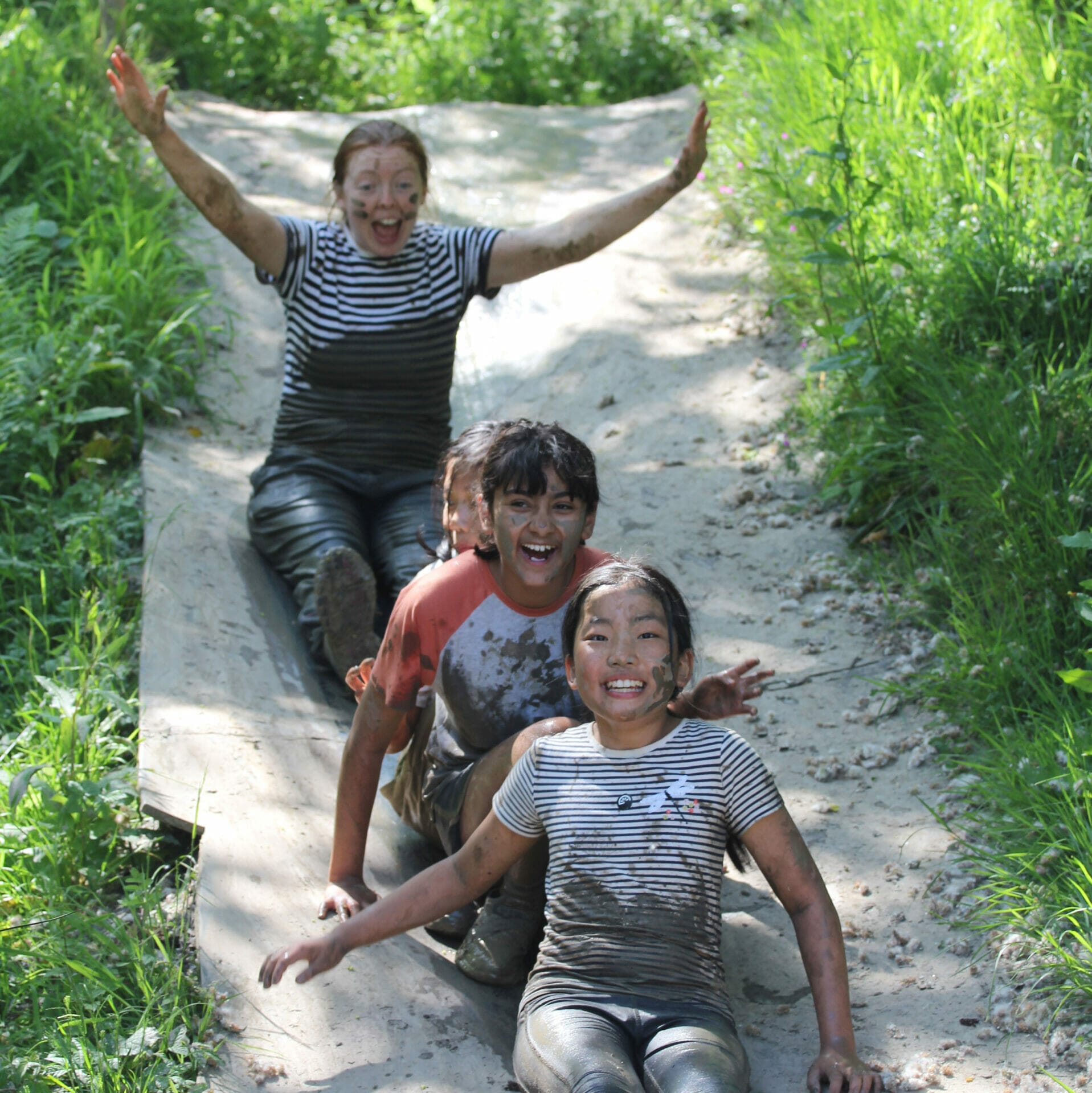 Three pupils and their teacher slide down a mud slide with their arms in the air.