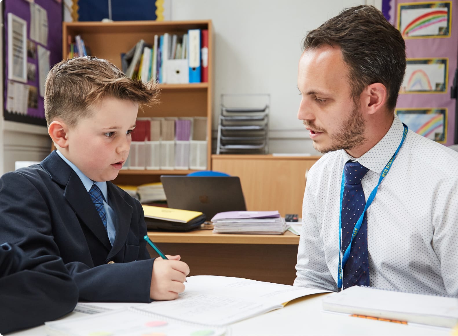 A teacher sat at a desk with a pupil who is writing and asking the teacher a question.