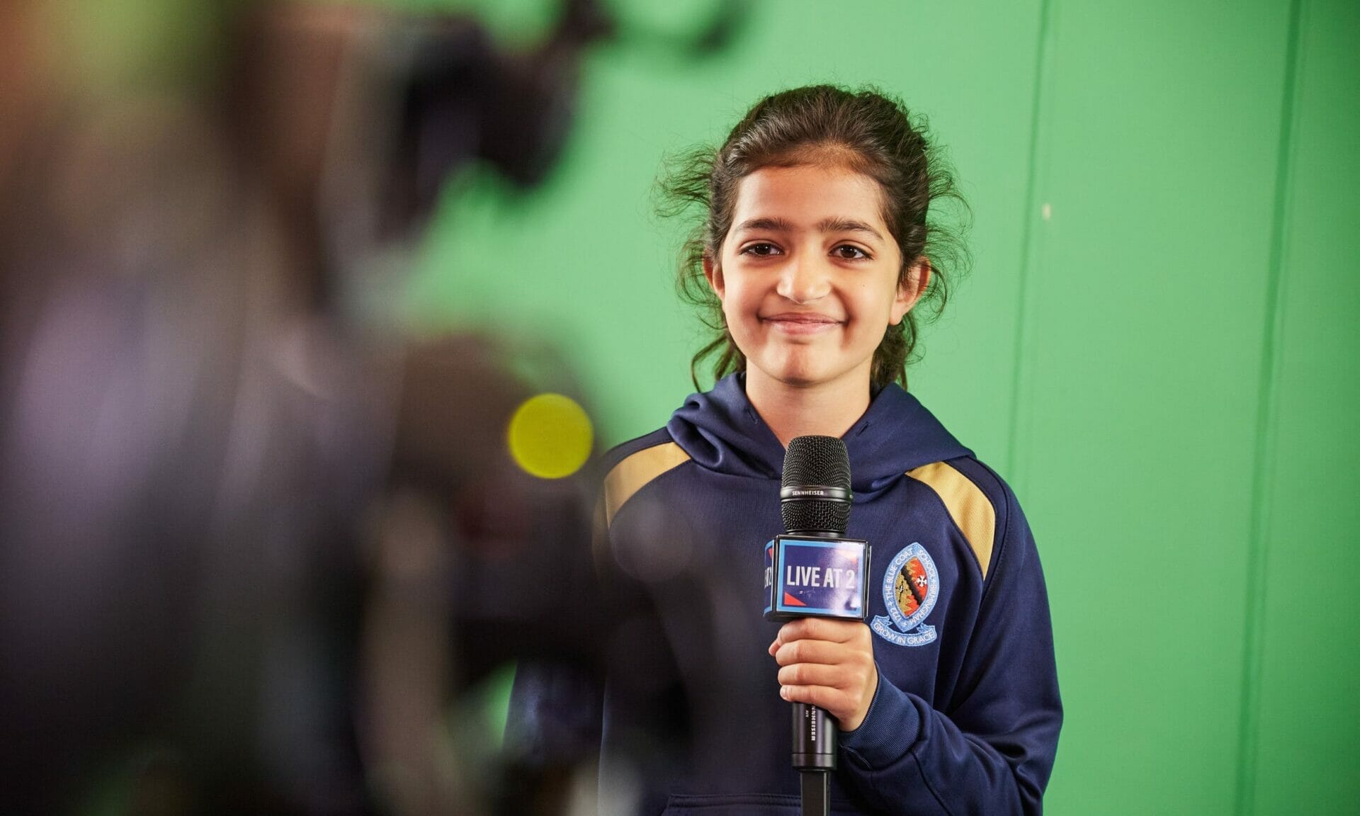A girl holds a microphone up in front of a green screen during a Computing lesson where they are learning how to broadcast.