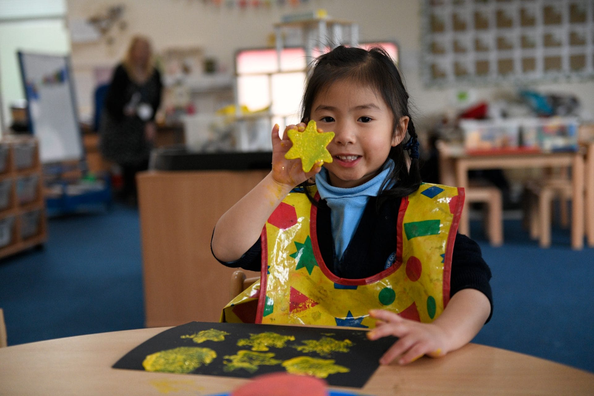 A girl holding up a star stencil with yellow paint on it and the piece of paper in front of her contains six yellow painted stars.