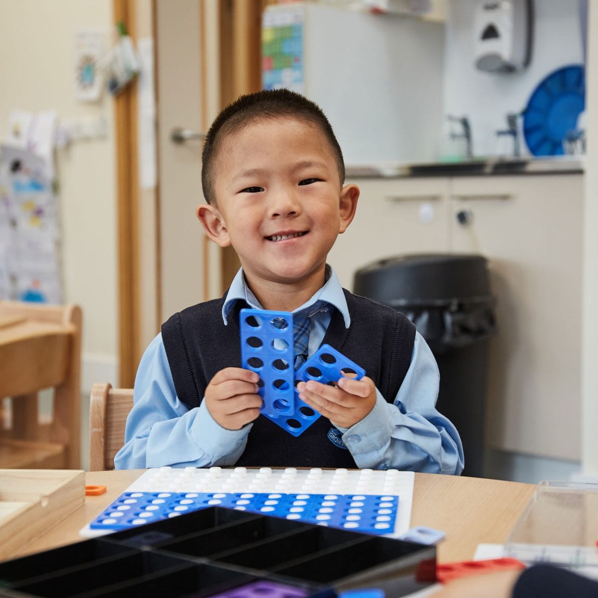 A boy in Nursery smiling at the camera holding a numicon shape.