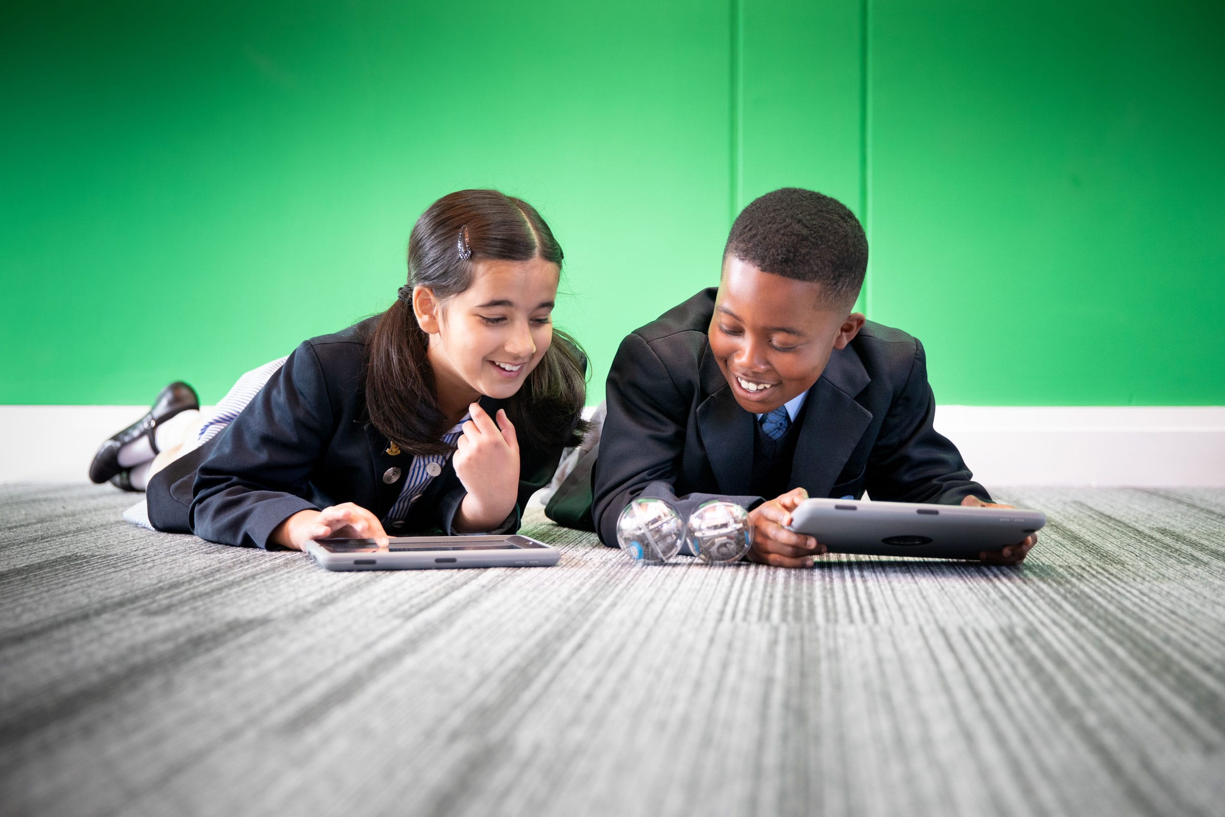 Two pupils laying on the floor looking and smiling at their iPads.
