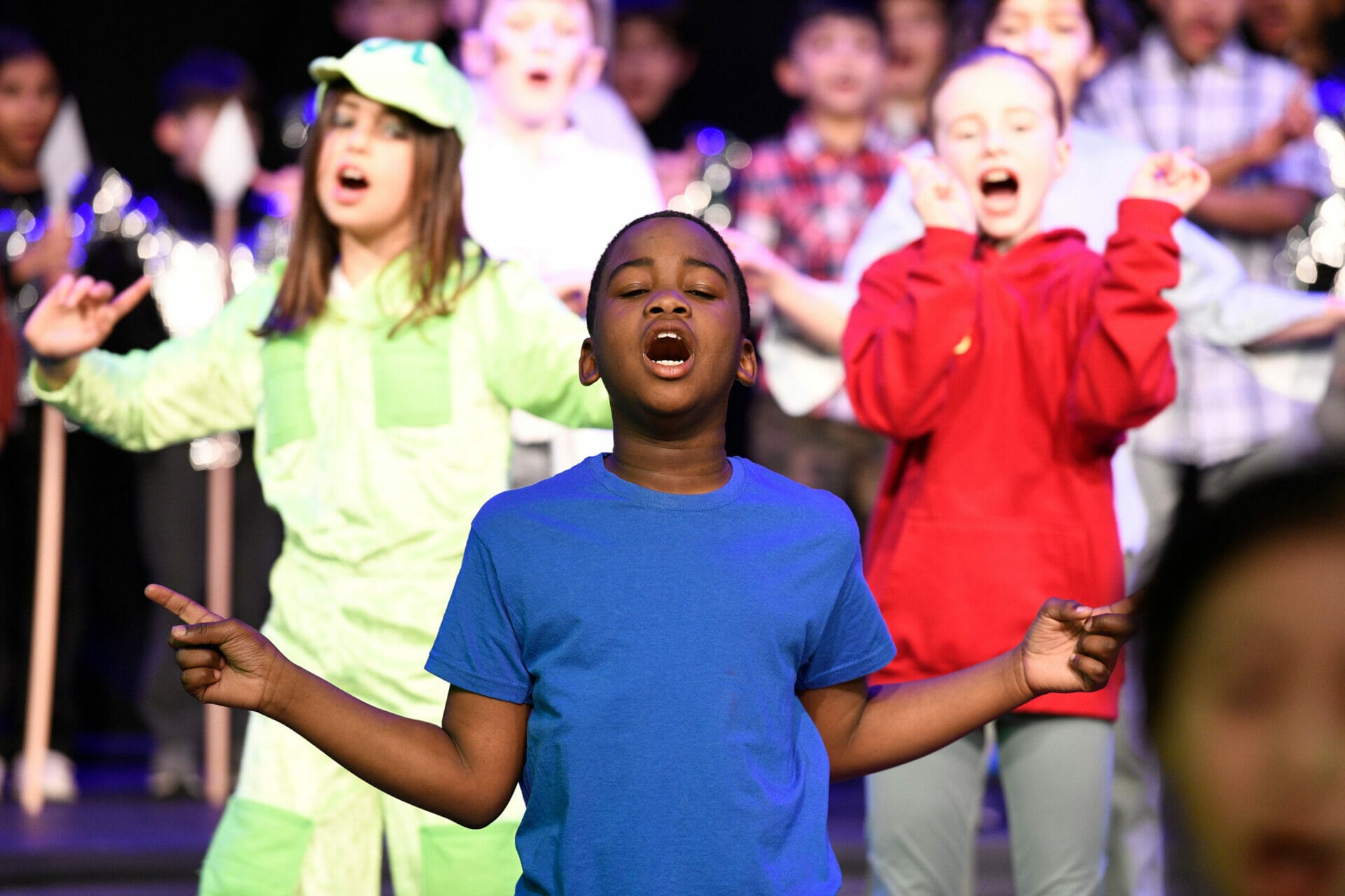 A boy in a blue t-shirt has his eyes closed and fingers pointing outwards whilst singing in a group.