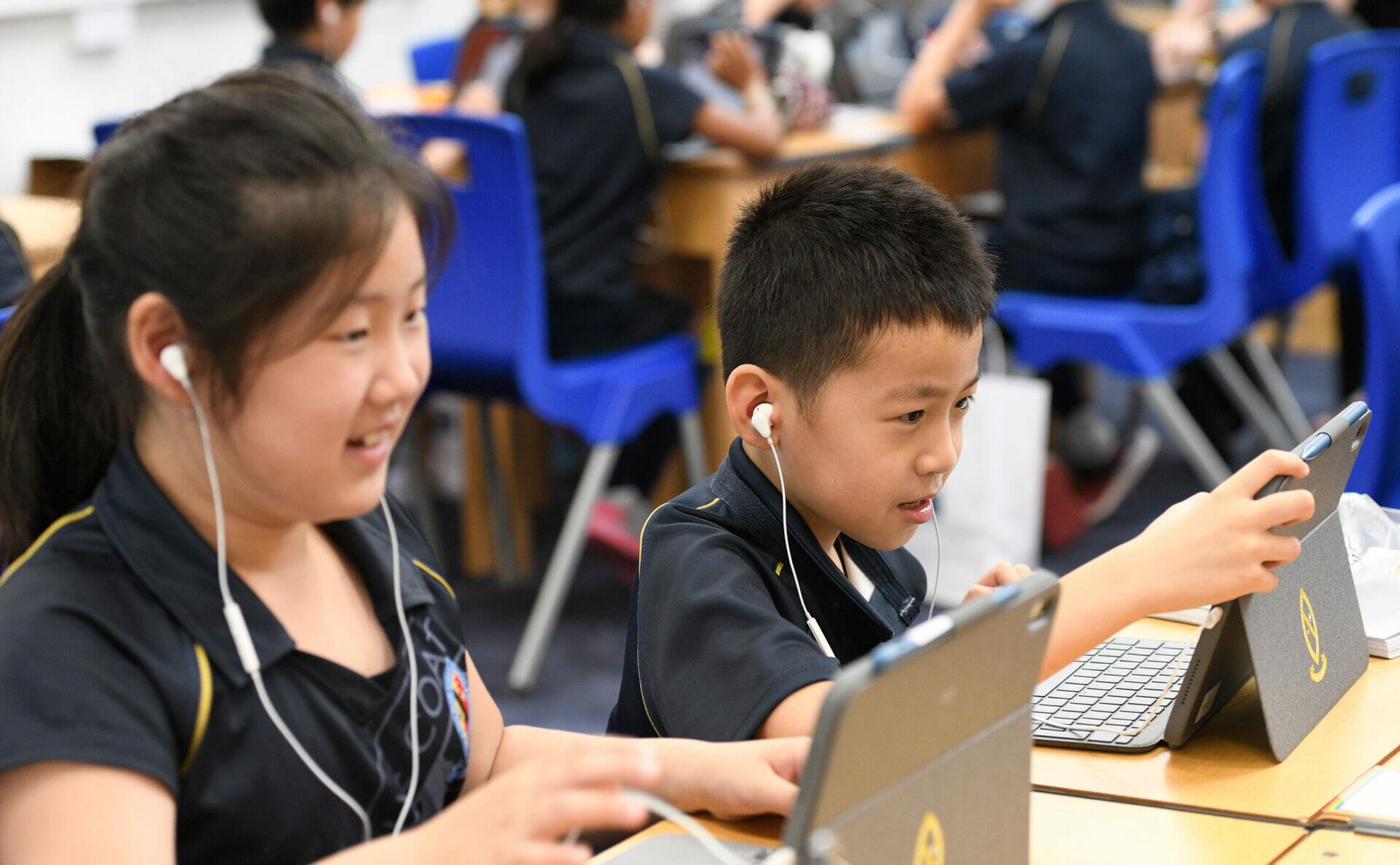 Two pupils sit at a desk working on their iPads with earphones in.